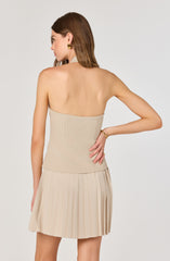 A woman is wearing a strapless, beige ribbed top and a matching pleated skirt in a light beige color. She has shoulder-length brown hair and is accessorized with gold earrings and a gold bracelet.