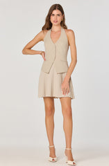 A woman is wearing a sleeveless beige vest with vertical pinstripes and a V-neckline, buttoned down the front, with pockets on each side. She pairs it with a matching beige pleated skirt that falls above the knees. She completes her outfit with white high-heeled sandals with ankle straps.