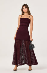 A young woman is wearing a strapless burgundy dress with thin spaghetti straps. The dress features textured fabric on the fitted bodice and a lace, semi-sheer skirt with a fitted lining from the waist up and a flowy, transparent overlay extending to the floor. She is carrying a small black handbag in her right hand and is wearing black high-heeled sandals with thin straps.