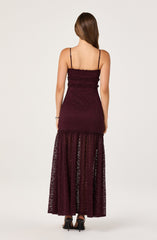 A woman is wearing a long, burgundy dress. The top portion of the dress is fitted with thin spaghetti straps and features horizontal ruffle accents. The lower part of the dress transitions into a sheer, lace fabric with vertical patterns, extending to the floor.