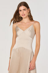 A woman is wearing a sleeveless dress with thin spaghetti straps. The top part of the dress is made of a shiny, satin-like fabric in a light beige or champagne color, featuring a V-neckline. The skirt portion of the dress is pleated, also in a matching light beige or champagne color, creating a textured, flowing appearance. She accessorizes with large, gold hoop earrings and a gold bracelet on her left wrist.