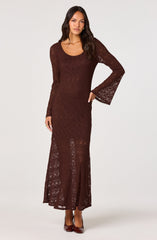 A woman is wearing a long, brown lace dress with a round neckline. The dress features long, semi-sheer bell sleeves and the lace pattern is intricate and see-through, revealing a slip underneath. The dress extends to the floor with a slight slit at the front. She is also wearing matching brown high-heeled shoes.