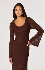 A woman wearing a fitted, long-sleeved, dark brown lace dress with a round neckline. The sleeves are bell-shaped, flaring out at the wrists. The dress has a sheer, intricate lace pattern throughout, revealing a slip underneath.