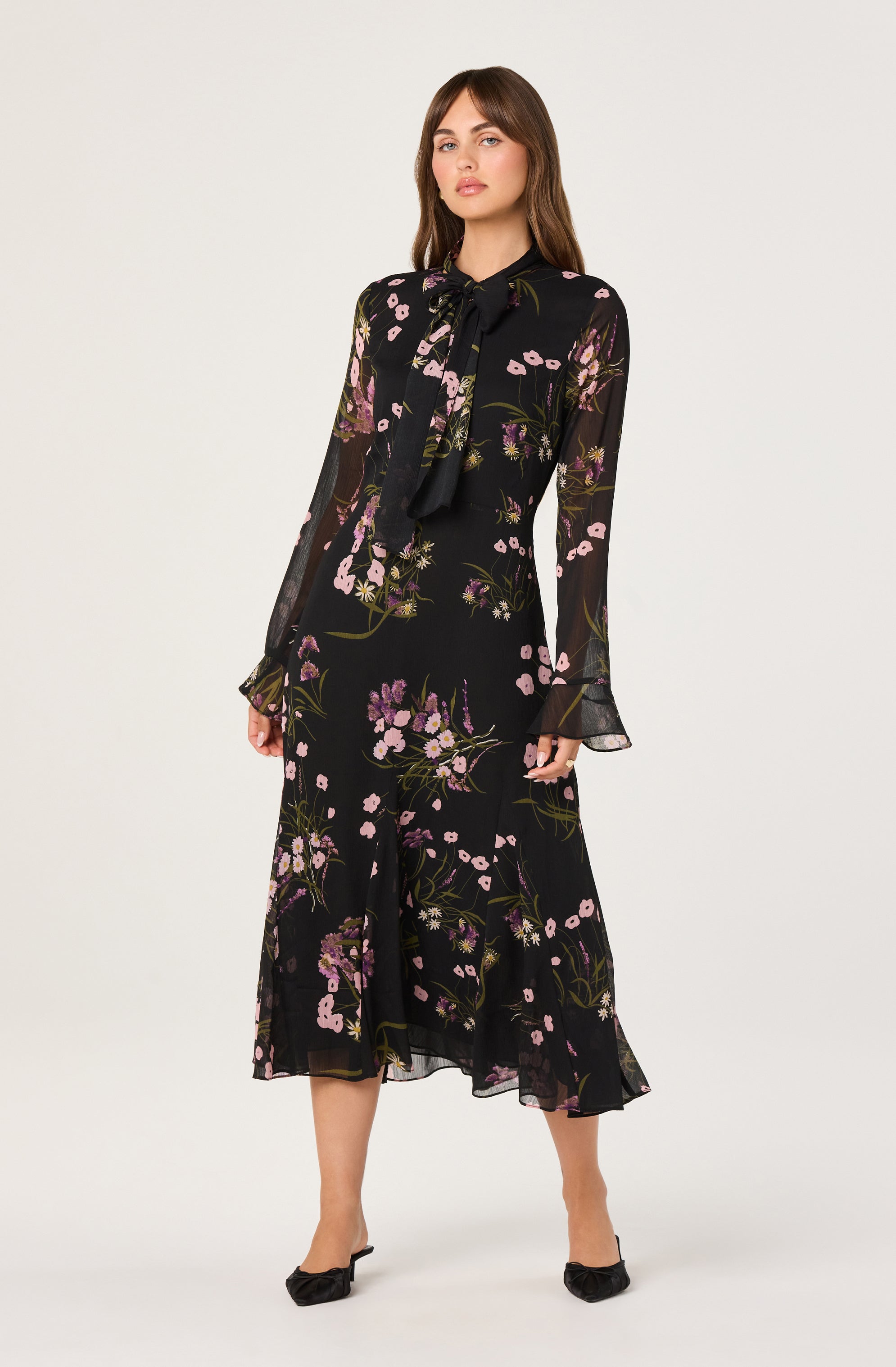 A woman is wearing a long black dress with a floral pattern of pink and purple flowers and green leaves. The dress has long, sheer sleeves with ruffled cuffs and a ruffled hemline that falls below the knees. She has a matching black tie bow at the collar.