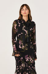 A woman is wearing a black long-sleeve dress with a floral pattern featuring pink, purple, and white flowers with green stems and leaves. The dress has a high neckline with a large bow tie at the front.