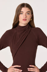 A woman is wearing a fitted, long-sleeve, high-neck dress in a deep brown color. The dress has asymmetrical draping detail across the front. She is accessorized with large gold hoop earrings and a gold ring on her right hand.