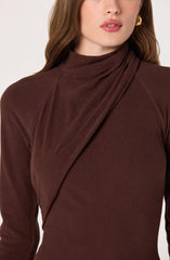 A woman is wearing a fitted, long-sleeve, high-neck top made of a smooth, dark brown fabric. The top features an asymmetrical draped detail across the chest and shoulder area. She accessorizes with a gold hoop earring.