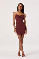 A woman is wearing a fitted, sleeveless, burgundy dress with spaghetti straps. The dress features decorative black bows on the chest area. She is also wearing strappy high-heeled sandals in a matching burgundy color, and gold jewelry including earrings and a bracelet.