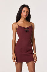 A woman is wearing a fitted, sleeveless, maroon mini dress with thin spaghetti straps. The dress features two decorative horizontal bows with embellishments on the front chest area.