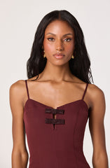 A woman with medium brown skin tone and long, dark wavy hair is wearing a burgundy sleeveless dress with thin spaghetti straps. The dress features decorative black beaded buckles in the front, creating two horizontal embellishments. She is also wearing gold hoop earrings.