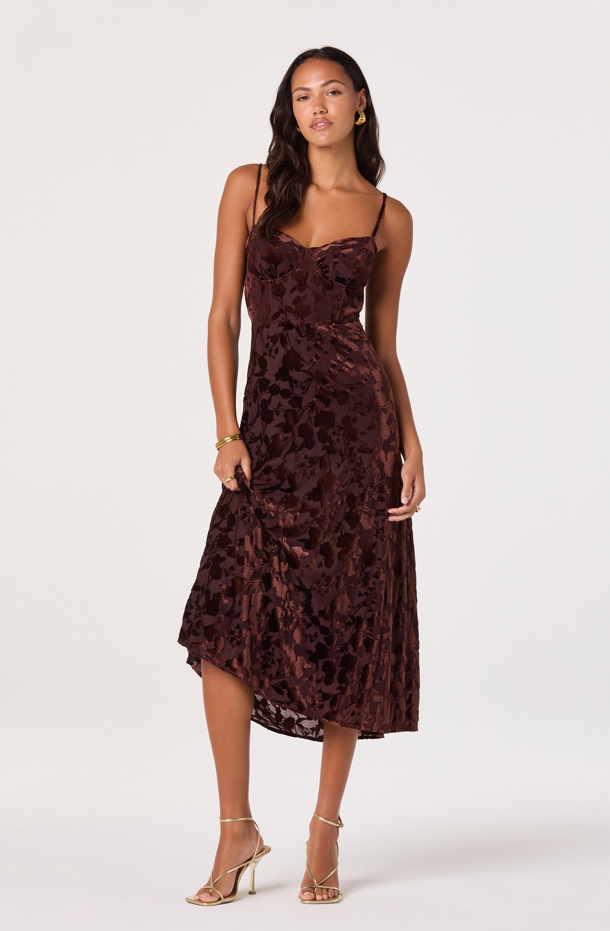 A woman is wearing a sleeveless, burgundy velvet dress with a floral pattern. The dress has thin spaghetti straps, a fitted bodice, and an asymmetrical hemline that falls below the knees. She is accessorized with gold hoop earrings, gold bracelets, and gold high-heeled sandals.