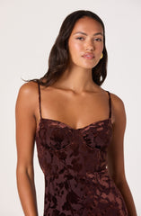 A woman is wearing a sleeveless, fitted dress with thin spaghetti straps. The dress has a floral pattern in shades of dark red and burgundy with a shiny, satin-like texture. She accessorized with gold earrings and has long, dark, wavy hair.