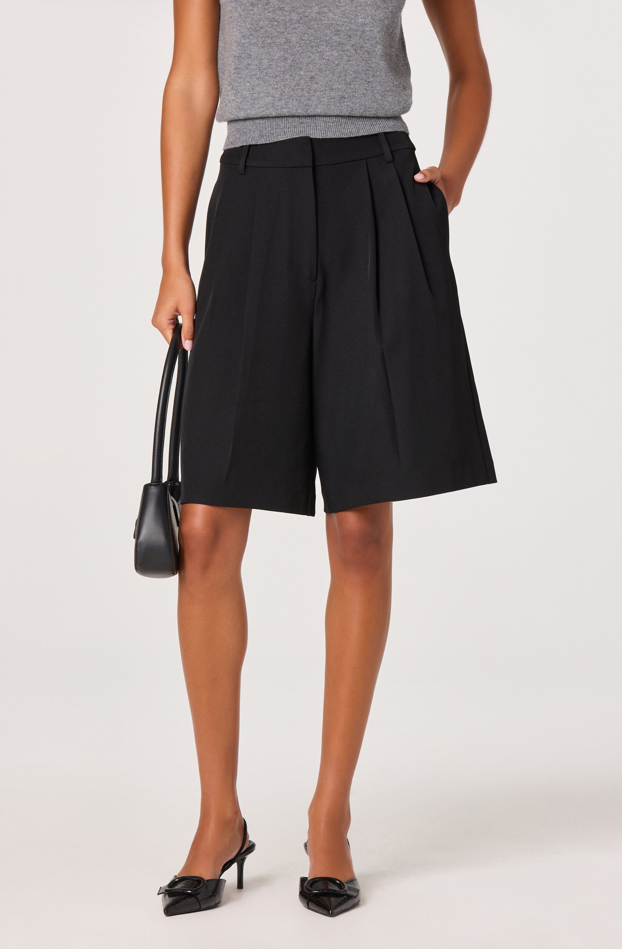 A person is wearing high-waisted black Bermuda shorts with wide legs and pleats, paired with a sleeveless gray top. They are holding a small black handbag in their right hand and are wearing black pointed-toe heels with bows on the front.