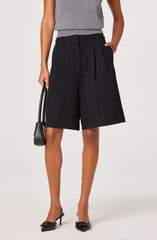 A person is wearing high-waisted black Bermuda shorts with wide legs and pleats, paired with a sleeveless gray top. They are holding a small black handbag in their right hand and are wearing black pointed-toe heels with bows on the front.