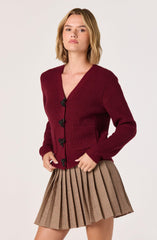 A woman is wearing a deep red, knitted cardigan with a textured pattern. The cardigan has a V-neckline and black decorative buttons shaped like bows down the front. She is also wearing a pleated skirt with a plaid pattern in shades of beige, brown, and black.