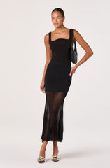 A woman is wearing a black sleeveless top with wide straps that has a fitted silhouette. She pairs it with a high-waisted black skirt that features a sheer, pleated overlay extending to the floor, creating a layered look with vertical stripes. She accessorizes with small gold earrings, a black shoulder bag, and open-toed high-heeled sandals with black straps and bow details.