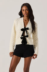 A woman is wearing a cream-colored, textured knit cardigan with long sleeves and a deep V neckline, accented by two large black bows at the front. She pairs it with black shorts made of a similar textured material.