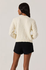 A woman is wearing a chunky, cream-colored knit sweater with textured and patterned sleeves, paired with high-waisted black knit shorts.
