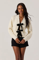 A woman is wearing a cream-colored knitted cardigan with a textured pattern, featuring two large black bows tied at the front. She pairs it with black shorts and has long, wavy dark hair.