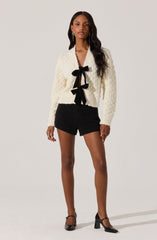 A woman is wearing a chunky, cream-colored knit cardigan with textured patterns, featuring a black ribbon-tied bow closure at the front. She pairs it with high-waisted black shorts and black pointed-toe heels with ankle straps.