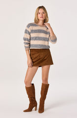 A young woman is wearing a long-sleeved, fuzzy sweater with wide horizontal stripes in beige and gray. She pairs it with a brown suede mini skirt and matching tall, heeled suede boots.