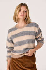 A woman is wearing a cozy, oversized sweater with wide horizontal stripes in neutral tones of beige and gray. The sweater has a round neckline and long sleeves. She is also wearing high-waisted, brown corduroy pants.
