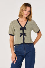 A woman is wearing a short-sleeved, knit cardigan with a black and white checkered pattern, featuring black trim on the V-neckline, sleeve cuffs, and hem. The cardigan has three decorative black bows down the front. She pairs it with high-waisted blue jeans.