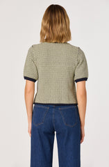 A woman is wearing a short-sleeved, textured beige and black patterned top with dark contrasting trim on the sleeves and hem, paired with high-waisted blue jeans. The back view shows the fit and design of the clothing.
