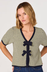 A woman is wearing a short-sleeved, V-neck sweater with a small, repeating geometric pattern in yellow and navy. The sweater features navy trim around the collar, sleeve cuffs, and down the front, accented with three matching navy bows along the front.