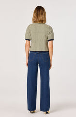 A person is wearing a short-sleeved, knitted sweater with a small checkered pattern in neutral tones and dark trim at the sleeves. They are also wearing high-waisted, wide-leg blue jeans. The outfit is casual, with the sweater and jeans paired together.