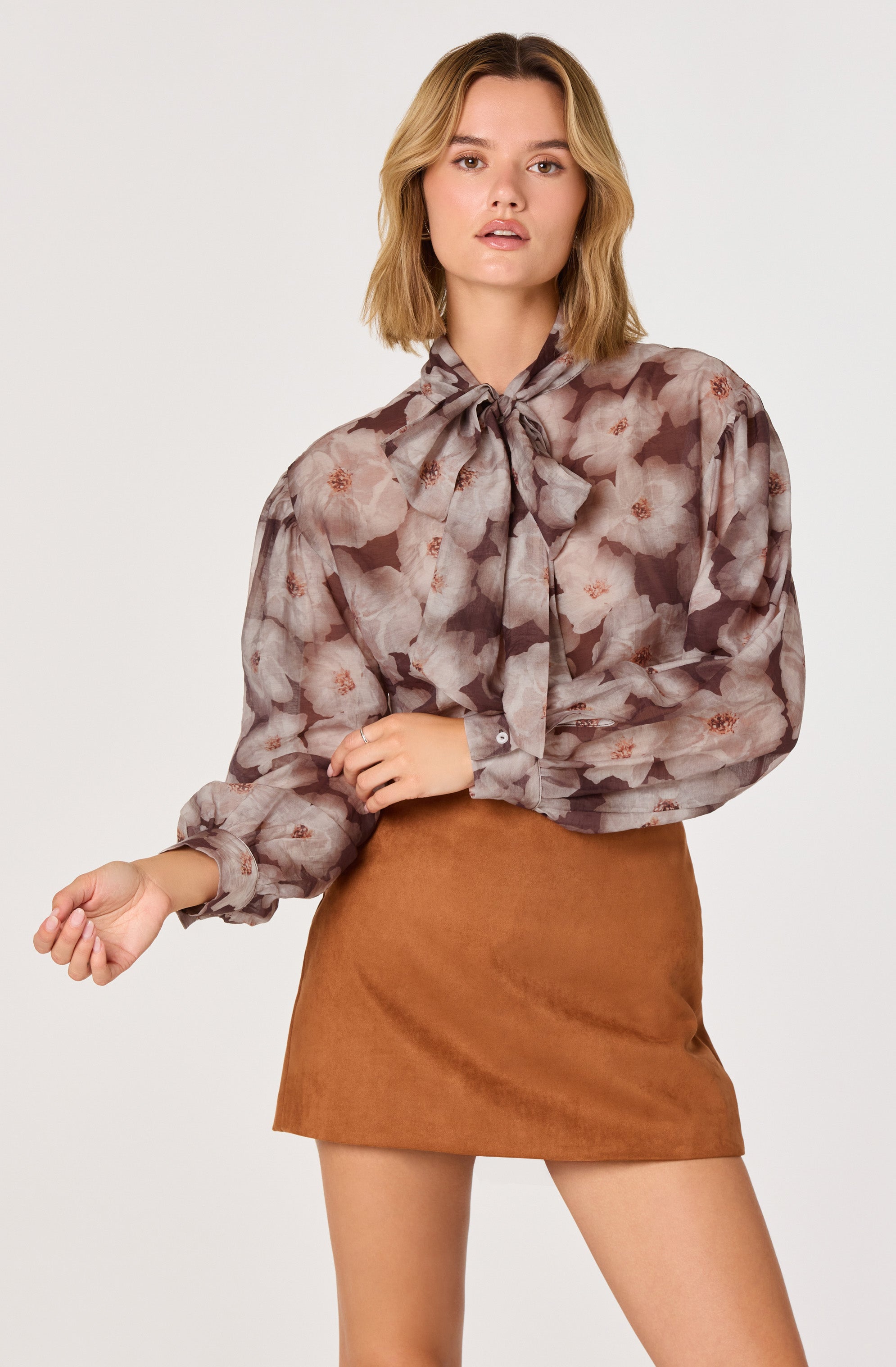 A woman is wearing a long-sleeved, semi-sheer blouse with a floral pattern in shades of pink, brown, and beige. The blouse features a large bow tied at the collar. She pairs this with a high-waisted, short brown skirt made of a suede-like material.