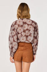 A woman is wearing a long-sleeved blouse with a large floral pattern featuring pale pink flowers on a semi-transparent, dark background. The blouse has a loose fit and gathered cuffs at the wrists. She pairs it with high-waisted, tan shorts made of a soft, suede-like material.