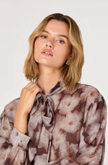 A woman wearing a long-sleeved, semi-sheer blouse with a floral pattern in shades of beige, brown, and blush pink. The blouse features a bow tie at the neckline and buttoned cuffs on the sleeves.