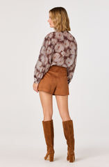 A woman is wearing a long-sleeved, loose-fitting blouse with a floral print in shades of pink, gray, and brown. She pairs it with high-waisted, brown suede shorts and tall, brown suede knee-high boots with chunky heels.