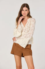 A woman is wearing a cream-colored lace blouse with long, slightly flared sleeves and a V-neckline with tie details. She pairs it with high-waisted, brown shorts made of a soft fabric. She accessorizes with gold hoop earrings and a ring on her left hand.
