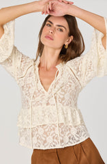 A woman is wearing a cream-colored, long-sleeved lace blouse with a floral pattern. The blouse features ruffles at the waist and a tie at the neckline. The fabric is semi-sheer, showcasing an intricate lace design with floral motifs. She is also wearing gold hoop earrings.