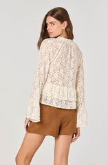 A woman is wearing a long-sleeved, cream-colored lace top with floral patterns, ruffle details at the bottom hem, and decorative stitching along the shoulders and upper back. She pairs the top with high-waisted brown shorts.