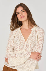 A woman with wavy brown hair is wearing a cream-colored, long-sleeved lace blouse with floral patterns and decorative openwork details along the shoulders. The blouse has a V-shaped neckline with a small ruffle trim. She is accessorized with gold hoop earrings and rings on her fingers.