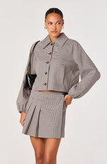 The outfit features a cropped, long-sleeve button-up jacket with a classic collar, made from a fabric with a small checkered pattern. The jacket has large, visible buttons and a chest pocket on the left side. The matching skirt is high-waisted with pleats on the front, also in a checkered pattern. The model accessorizes with gold hoop earrings and carries a small black shoulder bag with a gold accent strap.