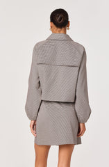 A woman is wearing a two-piece outfit consisting of a long-sleeved, tailored blazer with a collar and a matching high-waisted skirt. The blazer has a loose fit with slightly puffed sleeves, and the skirt is short, ending above the knees. The fabric features a small checkered or houndstooth pattern in neutral tones. The woman has her hair styled in a tight bun and is wearing small hoop earrings.