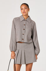 A woman is wearing a cropped, long-sleeved button-up shirt and a matching high-waisted pleated skirt. The shirt features a classic collar, a loose fit, and gathered elastic cuffs. The fabric has a small, checkered pattern in neutral tones, giving a sophisticated and timeless look. The skirt also has the same checkered pattern, with pleats that add texture and dimension to the outfit.