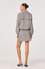 A woman is wearing a matching business suit consisting of a cropped blazer with a wide back yoke and long, elasticized sleeves, paired with a high-waisted, short A-line skirt. The fabric has a small checkered pattern in neutral tones. She completes the look with open-toe black high-heeled sandals.