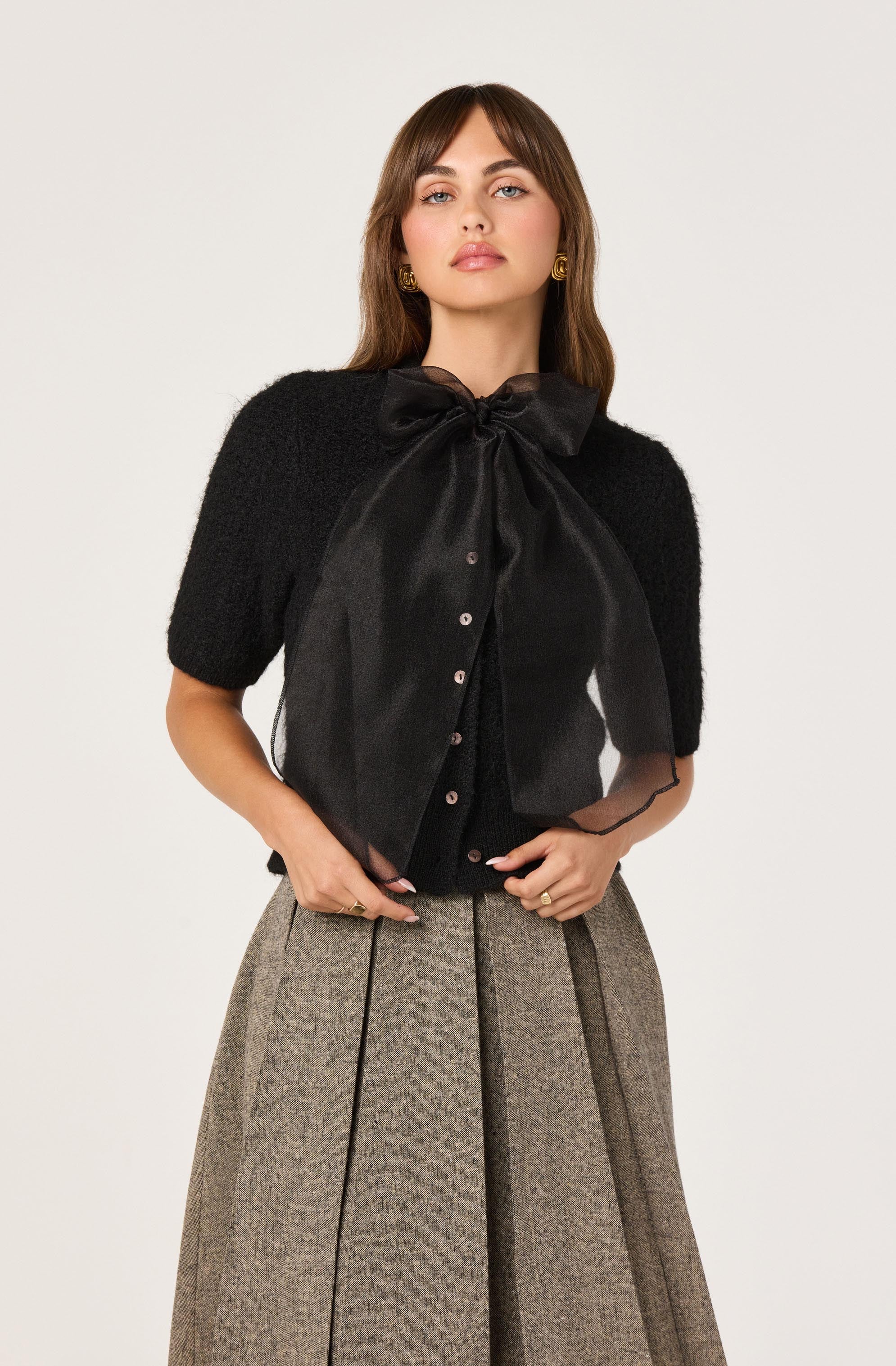 The woman is wearing a black short-sleeved cardigan made of textured, fuzzy material. Over it, she has a black satin blouse with a large bow at the collar and button closures down the front. She pairs this with high-waisted, wide-leg trousers in a light brown or taupe plaid pattern.