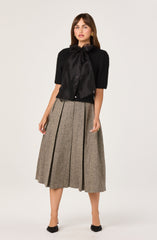 The woman is wearing a black short-sleeved sweater with a large satin bow tie at the neck. She has on a high-waisted, mid-length, pleated skirt made of a brownish tweed fabric with subtle black lines. She is also wearing black heels with a bow detail at the front.