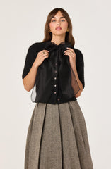 The woman is wearing a black short-sleeved, textured top, paired with a sheer black blouse that has a large bow at the collar and buttons down the front. She is also wearing a high-waisted, wide-leg skirt in a herringbone pattern featuring shades of gray and brown.