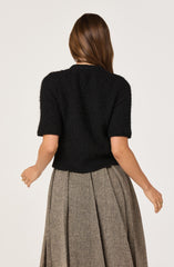 A woman is wearing a black, short-sleeved, fuzzy knit sweater and a high-waisted, pleated, gray plaid skirt.