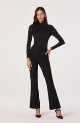 A woman is wearing a fitted black long-sleeve top with a high, ruched neckline, adorned with small white polka dots. She has on high-waisted black trousers with a straight-leg cut and subtle creases down the front. She is also wearing black pointed-toe high heels with ankle straps.