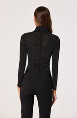 The image shows a woman from behind wearing a black, long-sleeved turtleneck top decorated with small, evenly spaced white dots. She is also wearing high-waisted black pants with belt loops and two back pockets. Her hair is brown and styled straight, falling past her shoulders.