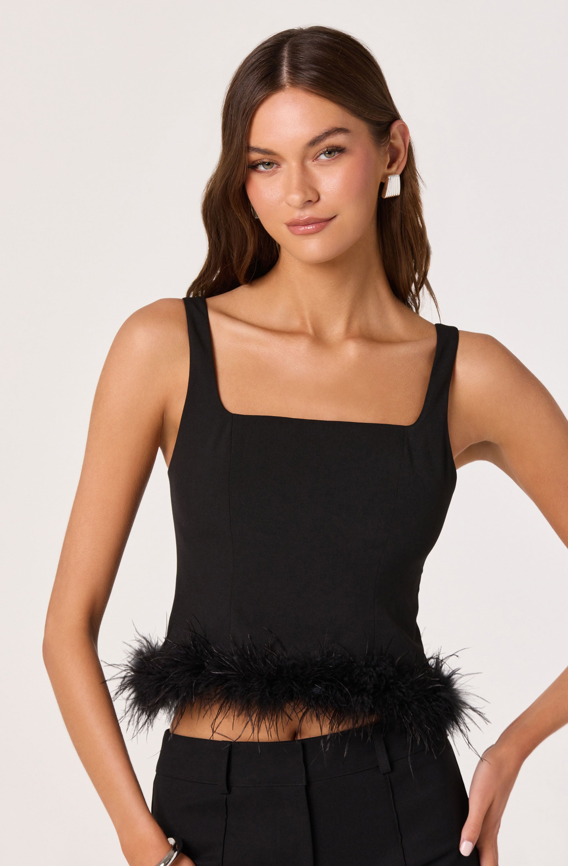 A woman is wearing a sleeveless black top with wide straps and a square neckline. The top features a decorative feather trim along the hemline. She is also wearing black pants and accessorized with white, rectangular statement earrings and a silver bracelet.