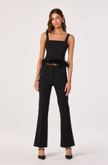 A woman is wearing a sleeveless black top with wide shoulder straps and a square neckline, featuring a feather trim at the hem. She pairs it with high-waisted, straight-leg black pants. She completes the outfit with black heels.